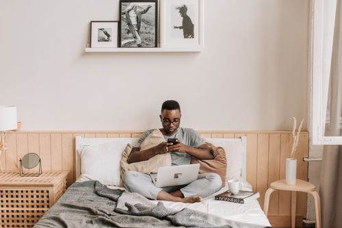 Free Person sitting on bed, using laptop and phone, in a bright cozy bedroom. Stock Photo