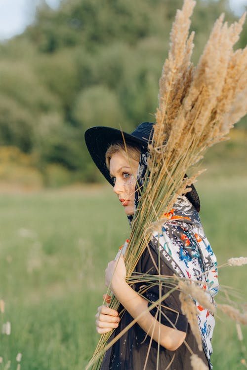 Free Woman in Blue and White Floral Dress Wearing Black Hat Holding Brown Woven Stick Stock Photo