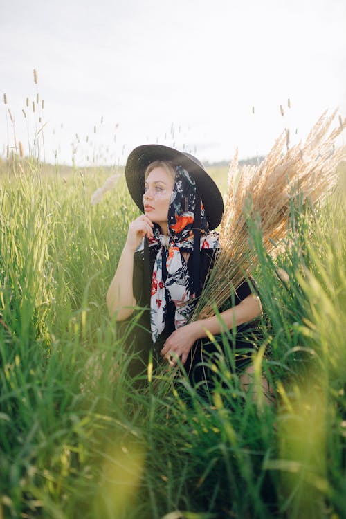 A Woman Wearing Black Sun Hat and Headscarf Sitting on Green Grass Field