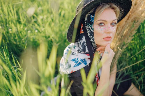 Woman in Floral Headscarf and Hat with Sparkles Under Eyes Sitting in Grass