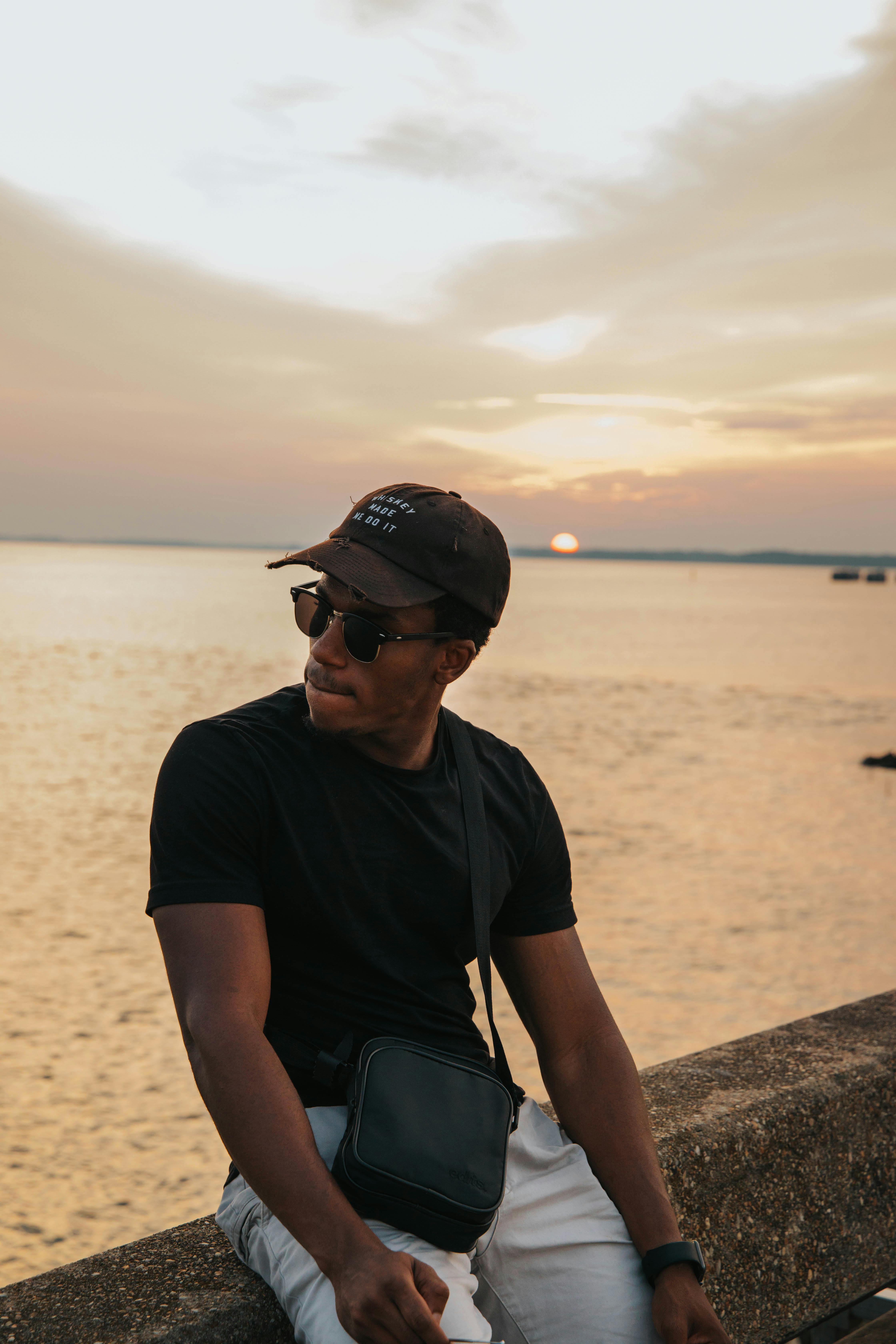 Young man in black attire sits by the water during sunset, wearing sunglasses and a cap.