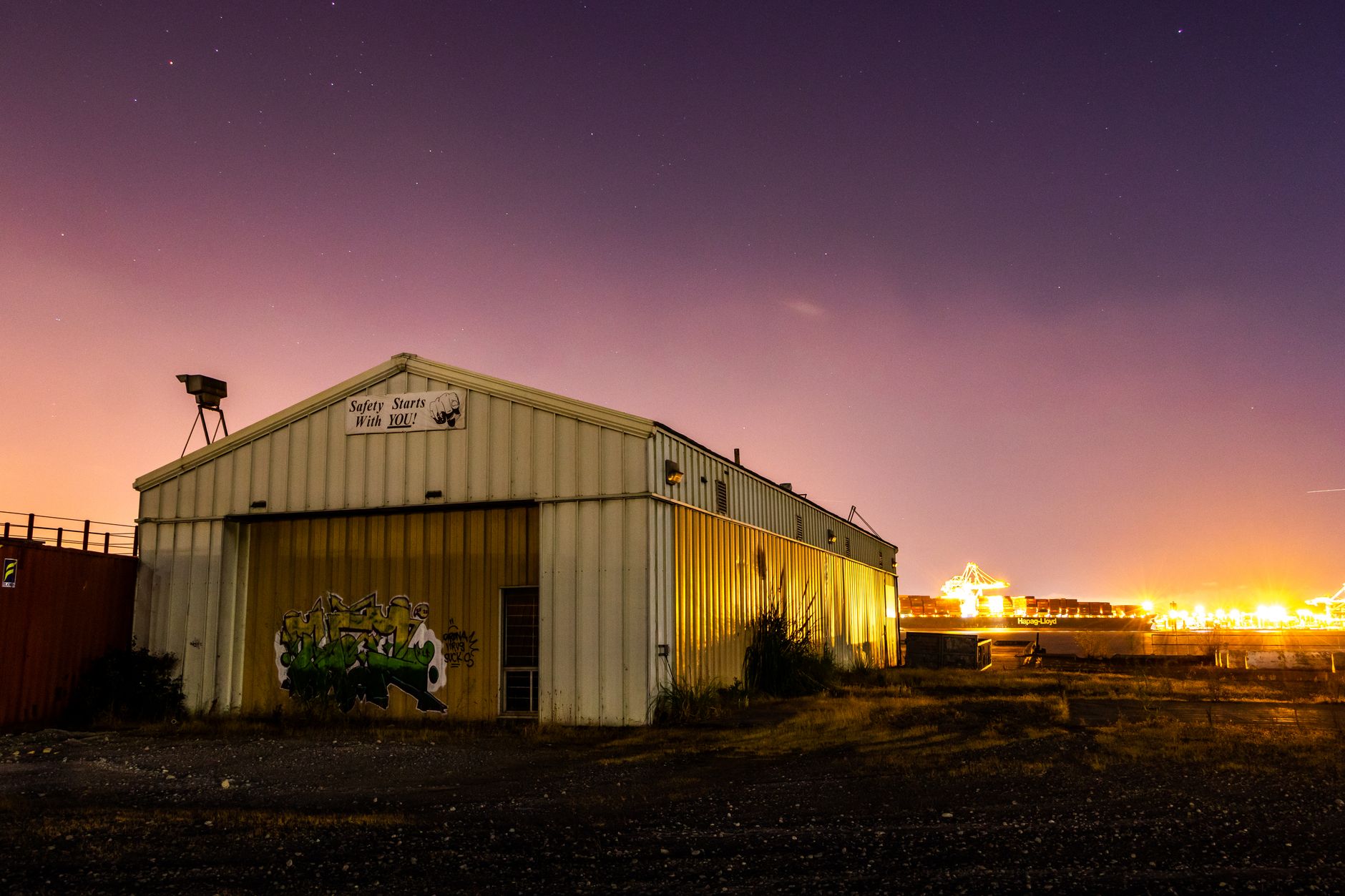 Nighttime rural warehouse entrance guarded by men while spectators gather around covered gamecock cages
