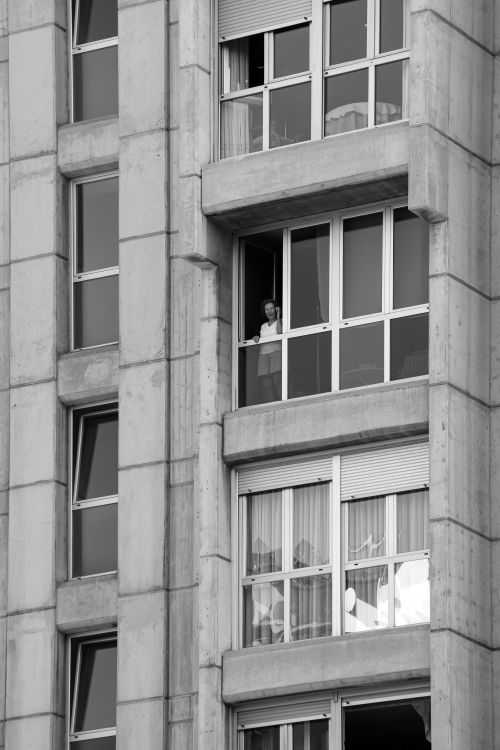 Free A person looking out from a high-rise building window in a cityscape. Urban lifestyle captured in monochrome. Stock Photo