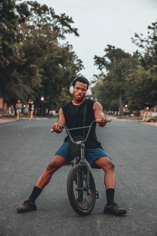Free A young man in a sleeveless shirt and headphones rides a bicycle outdoors on a tree-lined road. Stock Photo