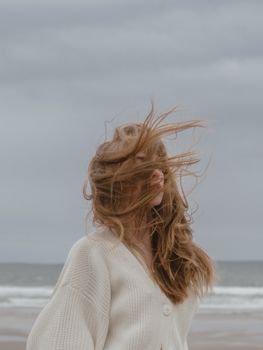 一位沉思的年轻女旅行者侧身而立，她留着长长的波浪卷发，身穿时尚针织羊毛衫，在阴云密布、海风轻拂的日子里，于海滩边悠然放松