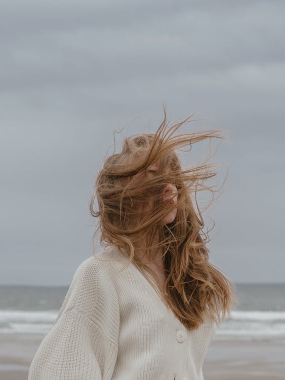 Side view of pensive young female traveler with long wavy hair in stylish knitted cardigan relaxing on beach of waving ocean against overcast sky on windy day