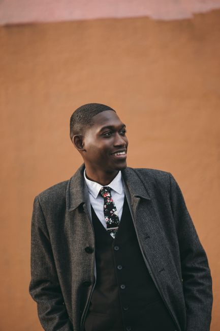 Confident young African American male model in elegant suit and coat standing against brown wall with hands in pockets and looking away