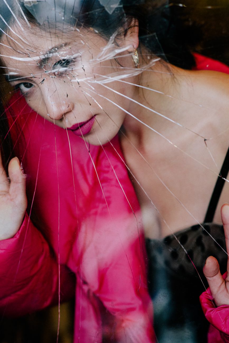 Through broken glass of crop young elegant Asian female with dark hair in fashionable outfit resting in room and looking