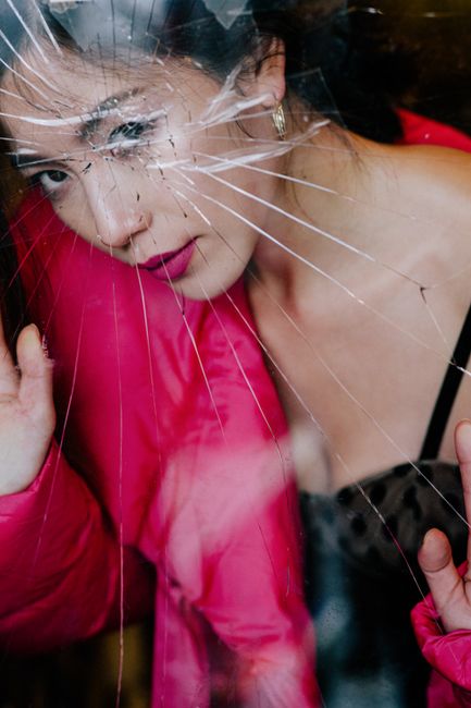 Through broken glass of crop young elegant Asian female with dark hair in fashionable outfit resting in room and looking at camera