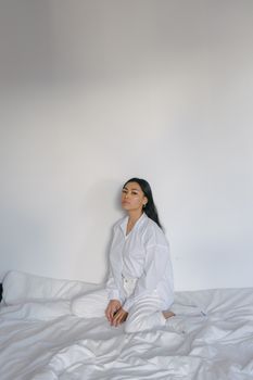 一位美丽的年轻少数族裔妇女侧身坐着，留着长长的黑发，身穿舒适的白色衣服，坐在柔软的床上，望着镜头