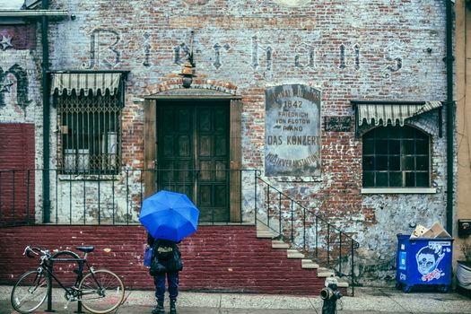 城市观光期间，一位身份不明的旅行者身着保暖衣物、手持雨伞，背对镜头站在古老砖砌建筑旁的街道上