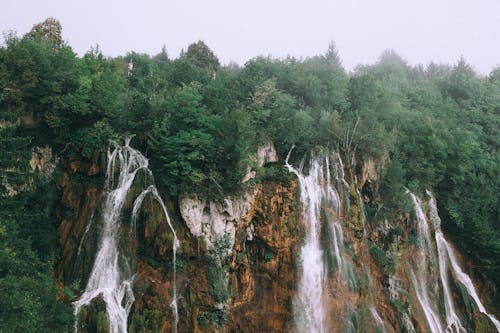 From below of picturesque rapid waterfall flowing among rough rocky cliff covered with lush green trees against cloudy sky
