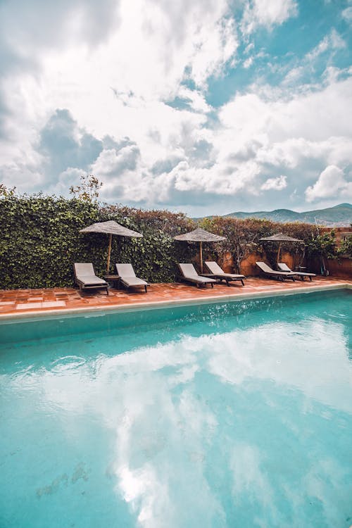 Peaceful scenery of deckchairs under umbrellas on poolside surrounded with green fence beneath blue sky in sunny resort