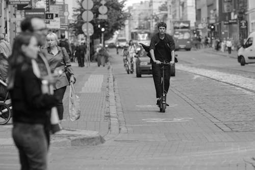 Black and white passers by walking and riding scooter along busy city road with moving cars on summer day