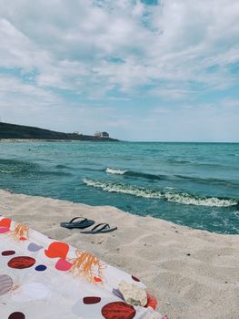 Amazing view of turquoise sea waving over sandy shore with flip flops and colorful beach towel on warm summer day
