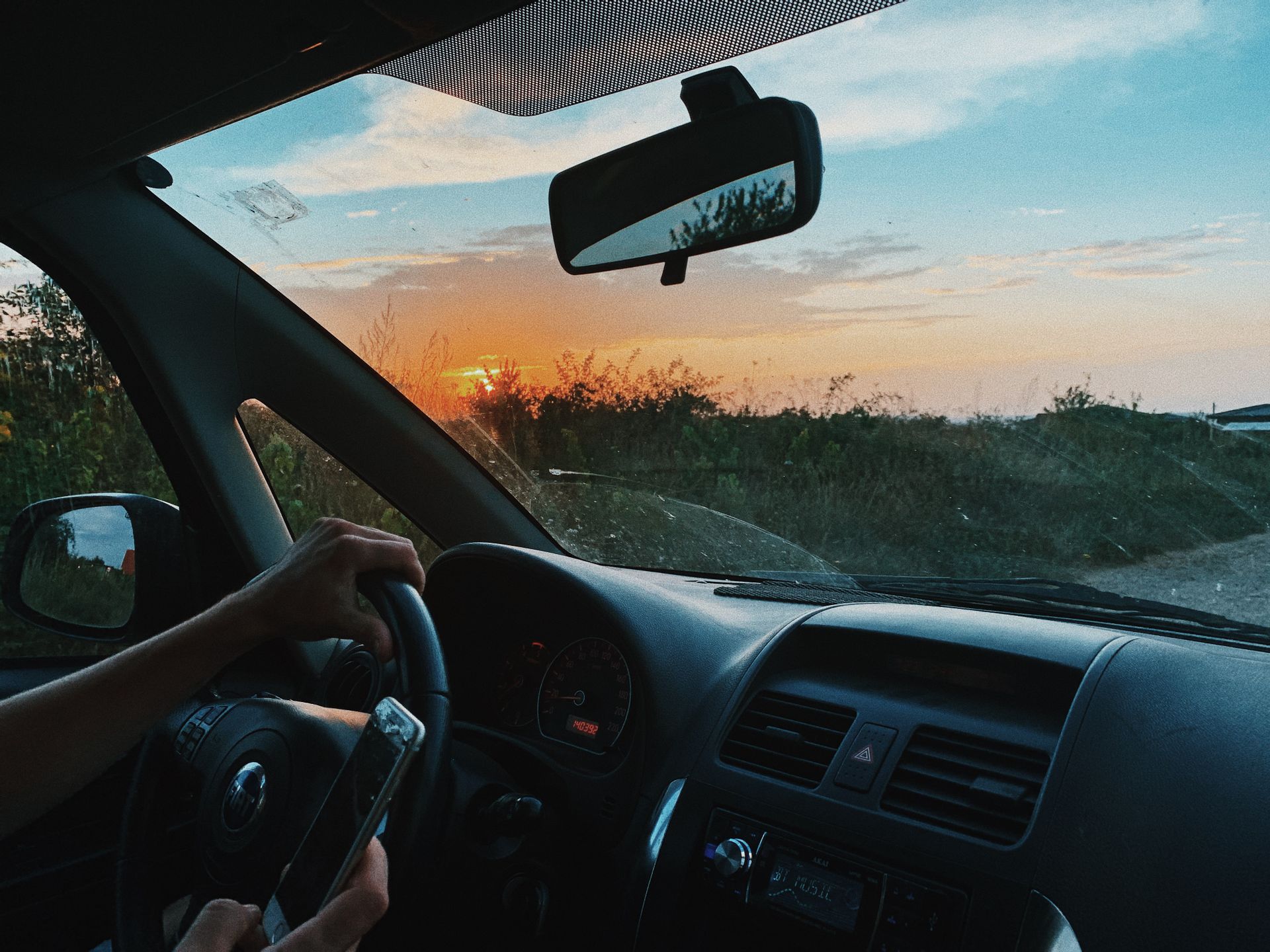 Crop unrecognizable person browsing mobile phone and driving car along rural gravel road in sunset time