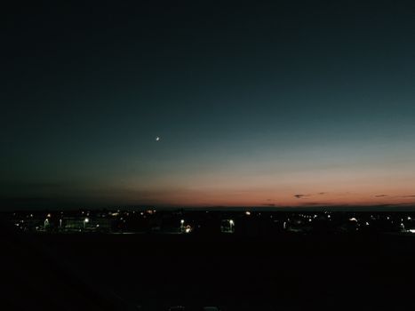 Starless dark sky with new moon glowing over small settlement houses illuminated by electric lights