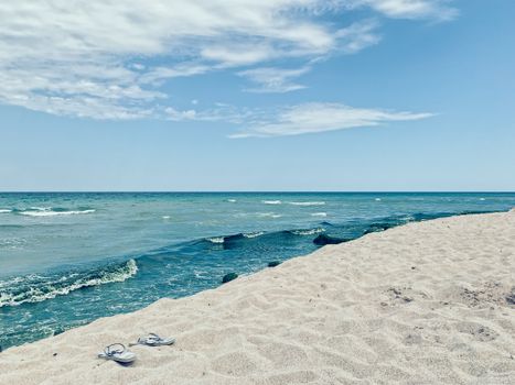 Small flip flops on sandy shore of vibrant dark blue wavy ocean under sky covered with clouds