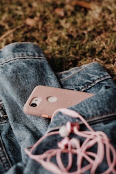 A close-up of a pink mobile phone and earphones on a denim jacket outdoors.