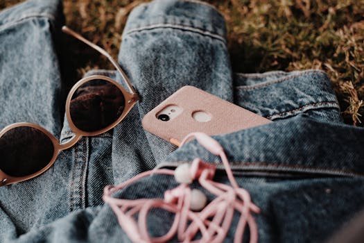 Close-up of sunglasses, phone, earphones, and denim jacket on grass, evoking casual vibes.