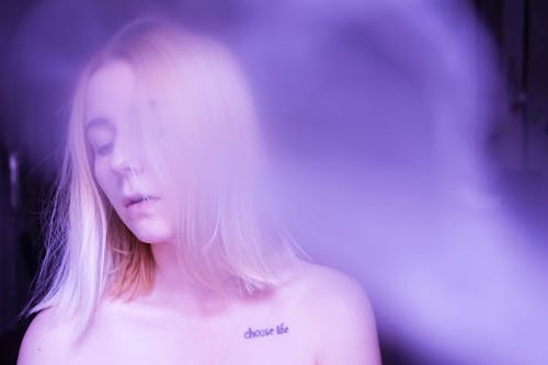 Free Pretty young female model with long blond hair and collarbone tattoo standing in smoke cloud in purple and blue light Stock Photo