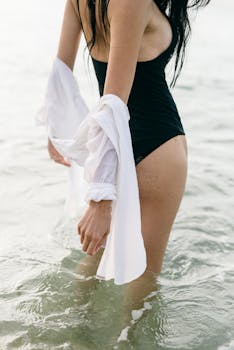 A woman in a black swimsuit wading through ocean water, showcasing summer fashion.