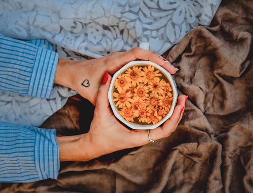 Top view of crop anonymous female warming hands by ceramic mug with hot beverage decorated with fresh flowers