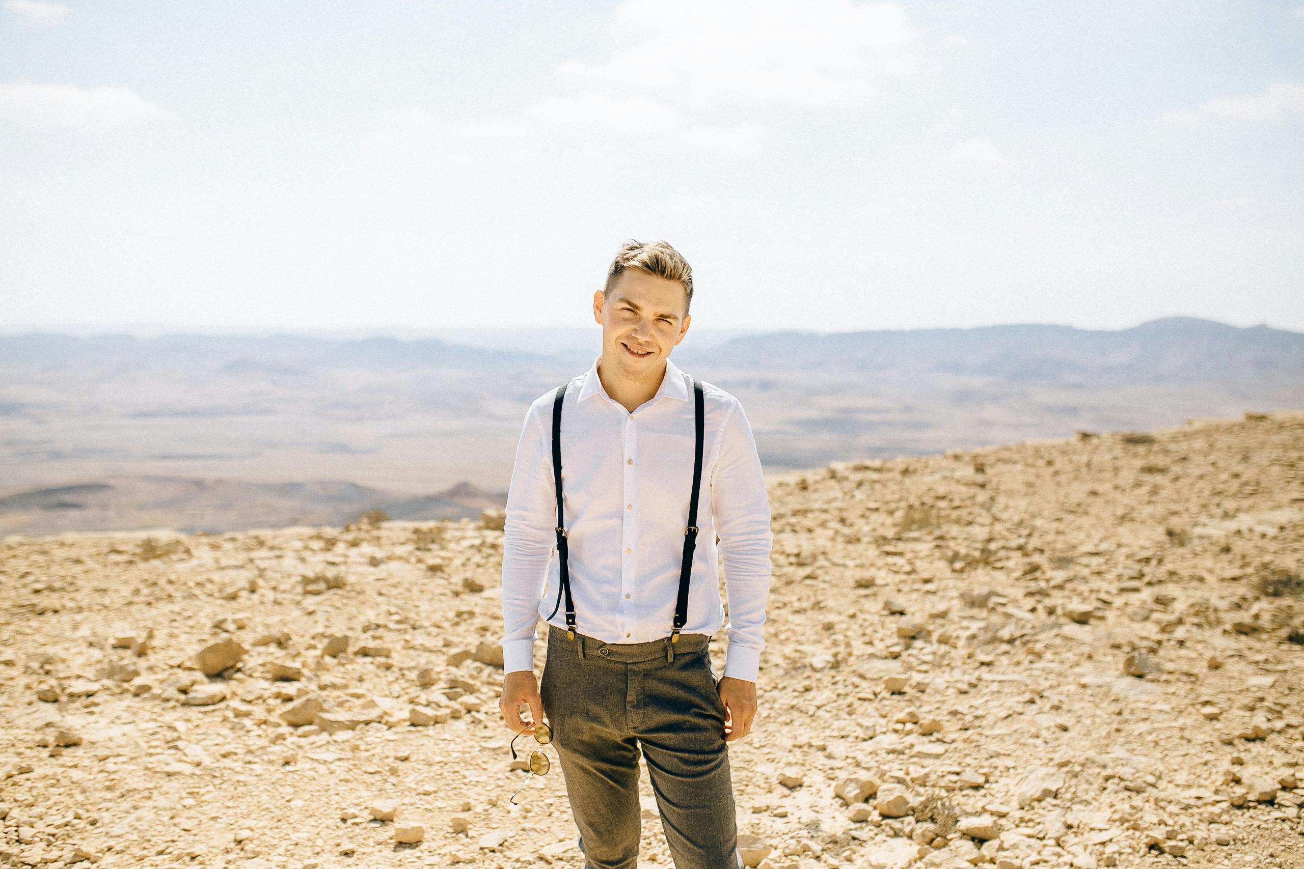 一名男子身穿白衬衫、系着背带，微笑着站在阳光明媚的沙漠中
