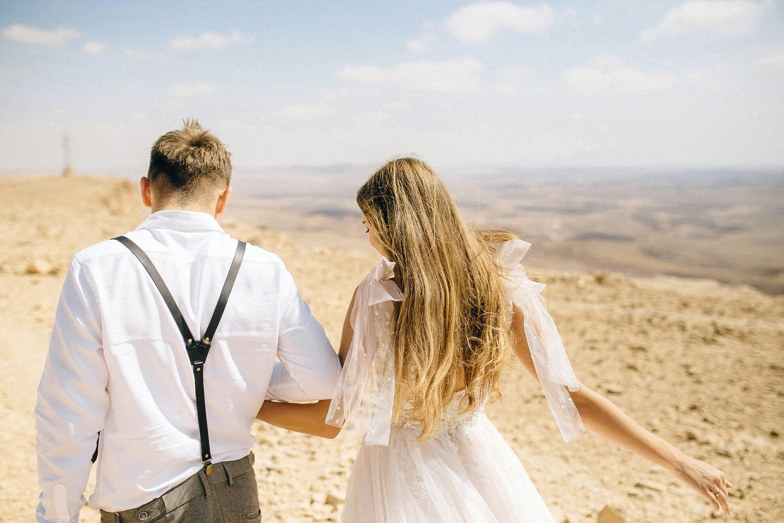 A couple in wedding attire walking hand in hand in a scenic desert setting, emphasizing love and adventure.