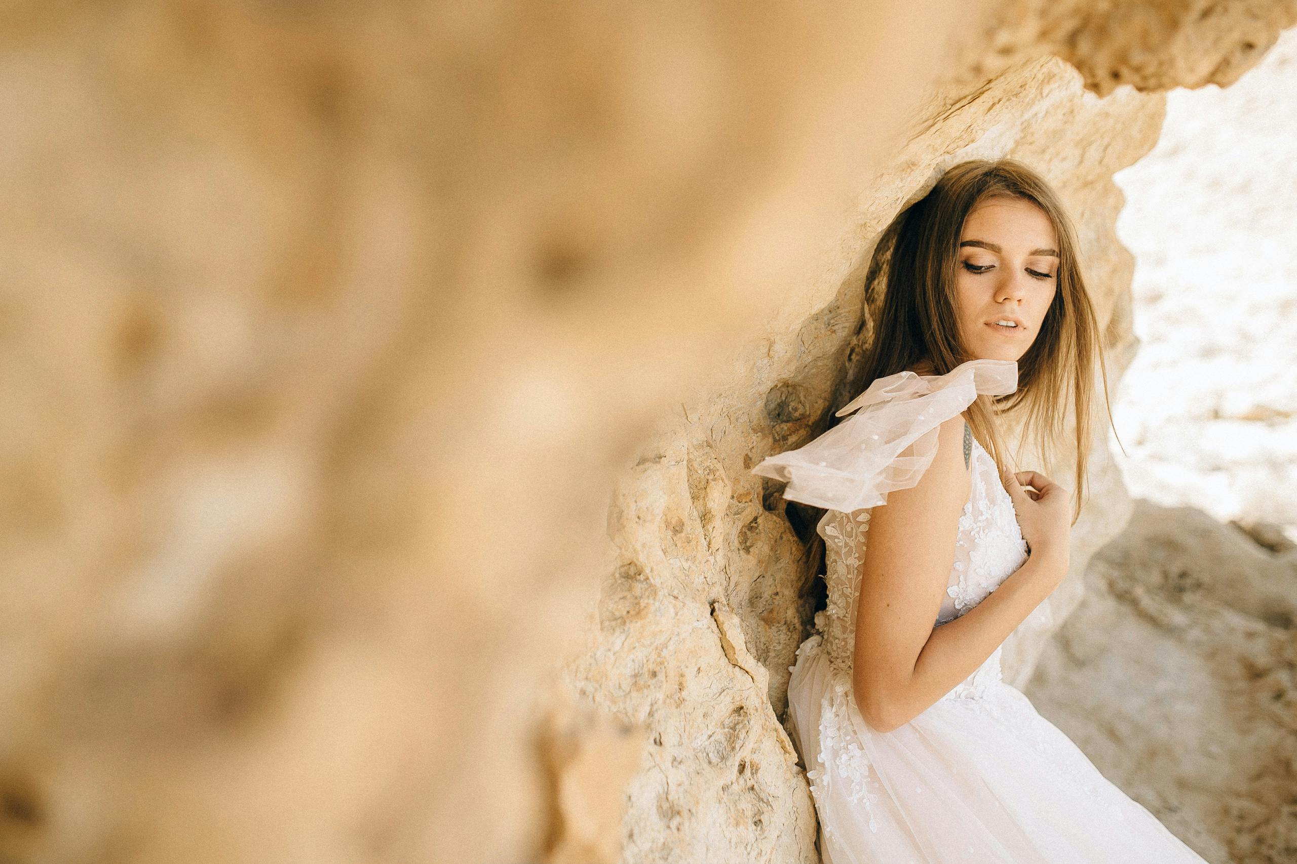 一位身着飘逸白裙的新娘，优雅地倚在质朴的石墙上摆着姿势，显得宁静而美好