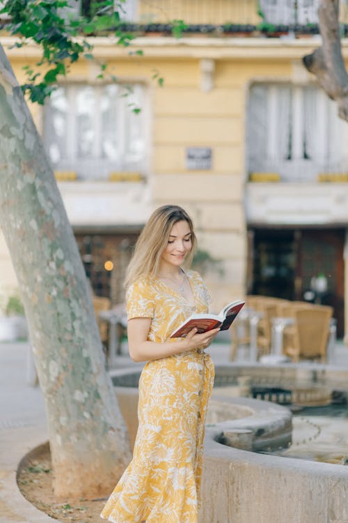 Woman in Yellow Floral Dress Standing while Reading Book