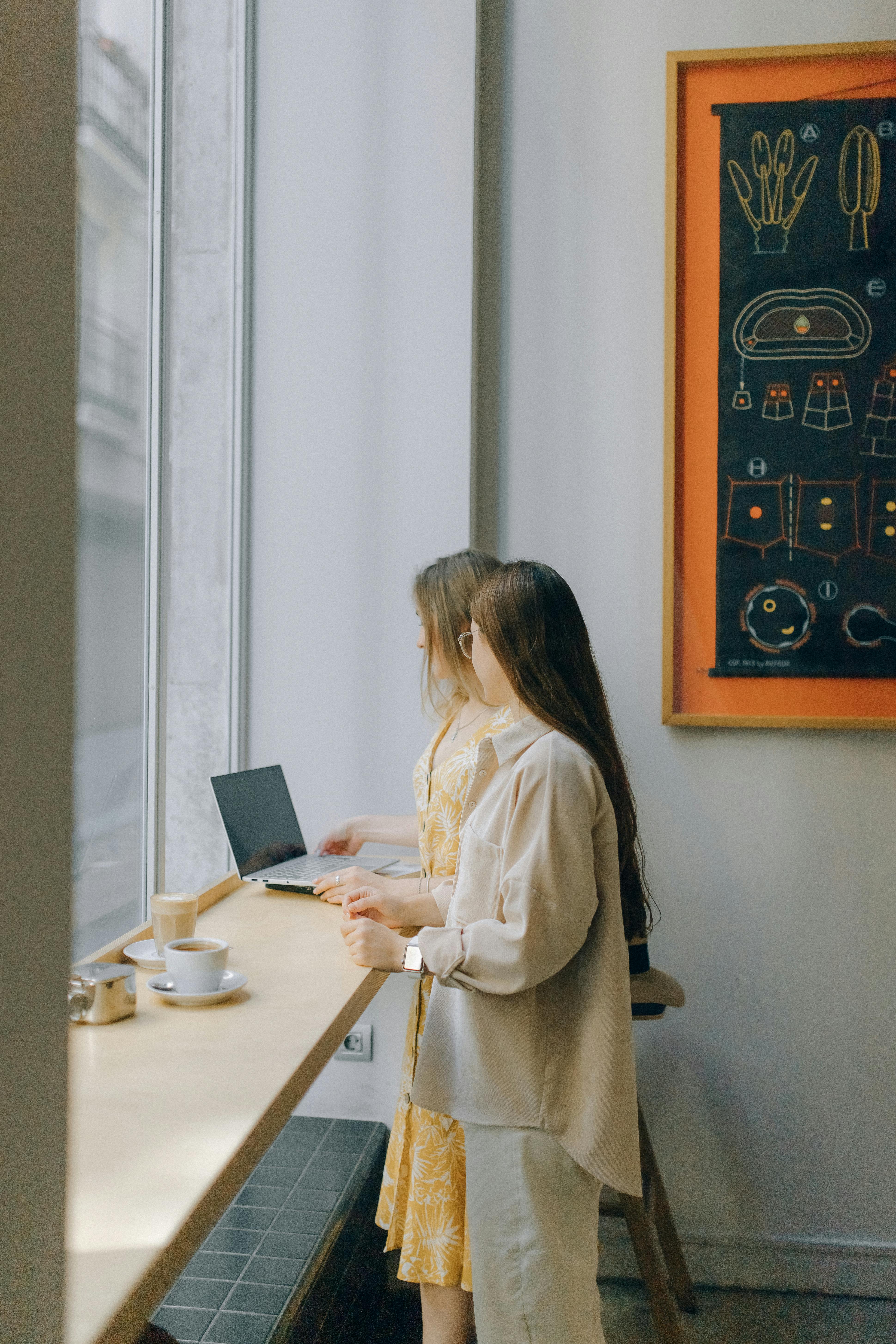 两名妇女站在窗前，用笔记本电脑做着室内休闲工作