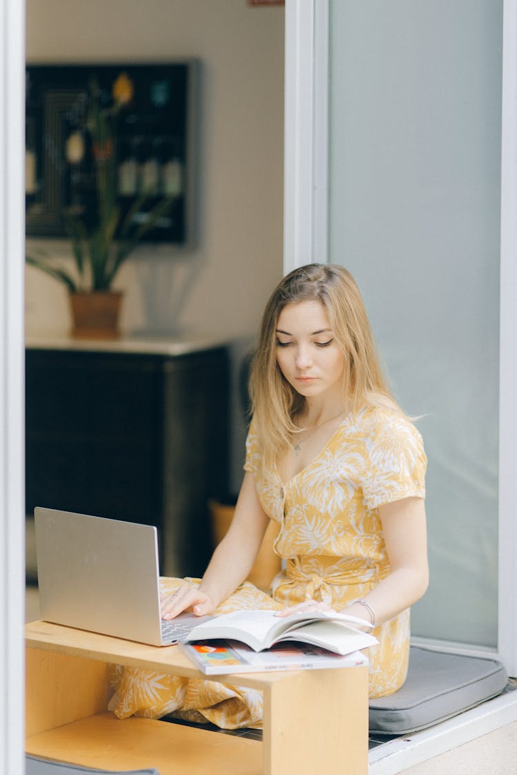 Woman In Yellow Dress Reading A Book While Using A Laptop