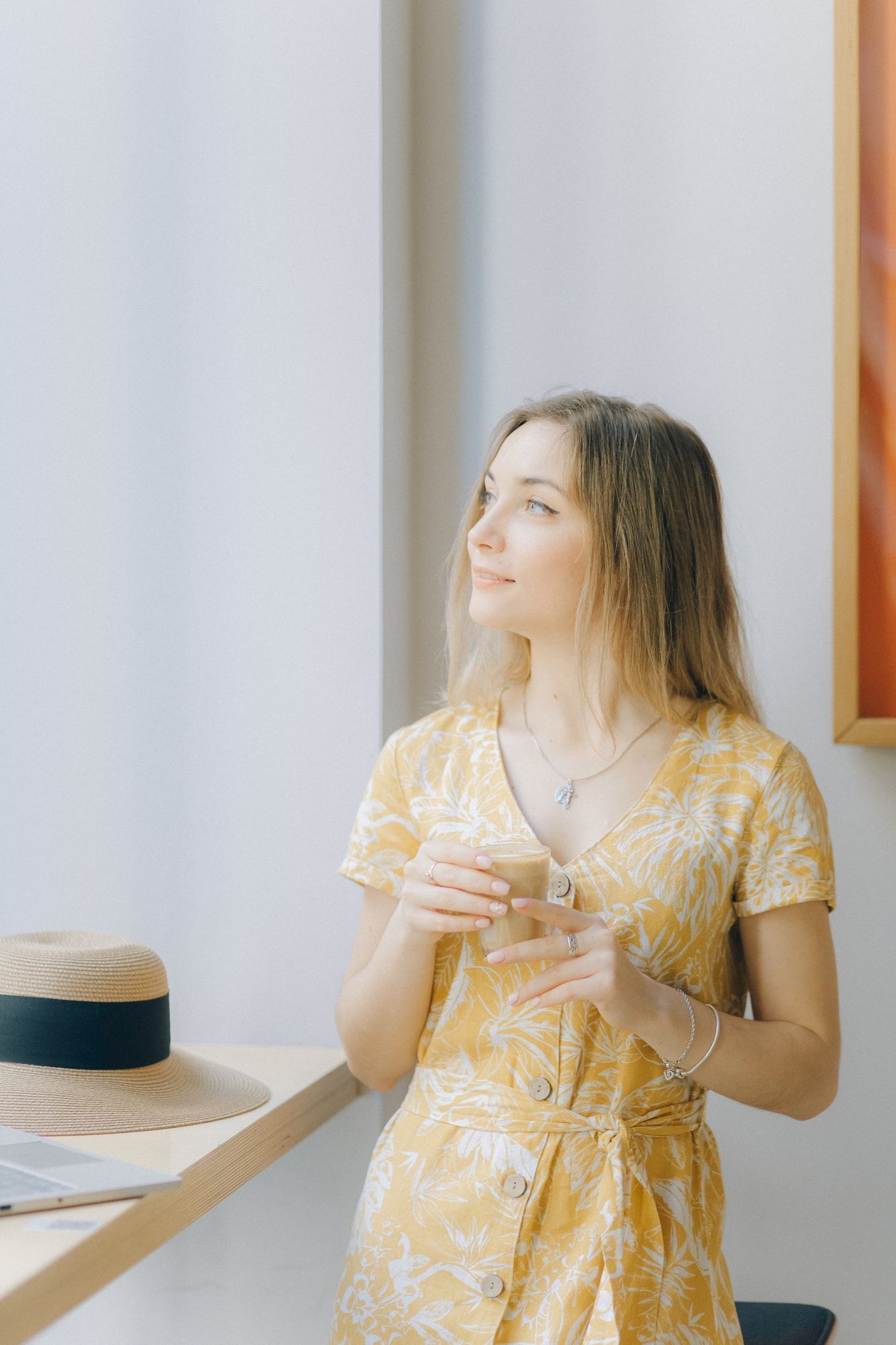 Woman in a yellow floral midi dress holding a coffee cup by a bright window