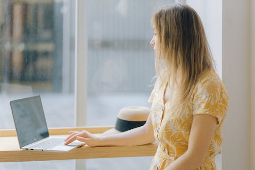 Woman in a bright yellow floral dress working remotely on a laptop by a window.