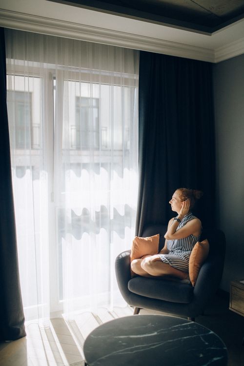Free A woman in a striped dress sits by a large window, basking in the natural light. Stock Photo