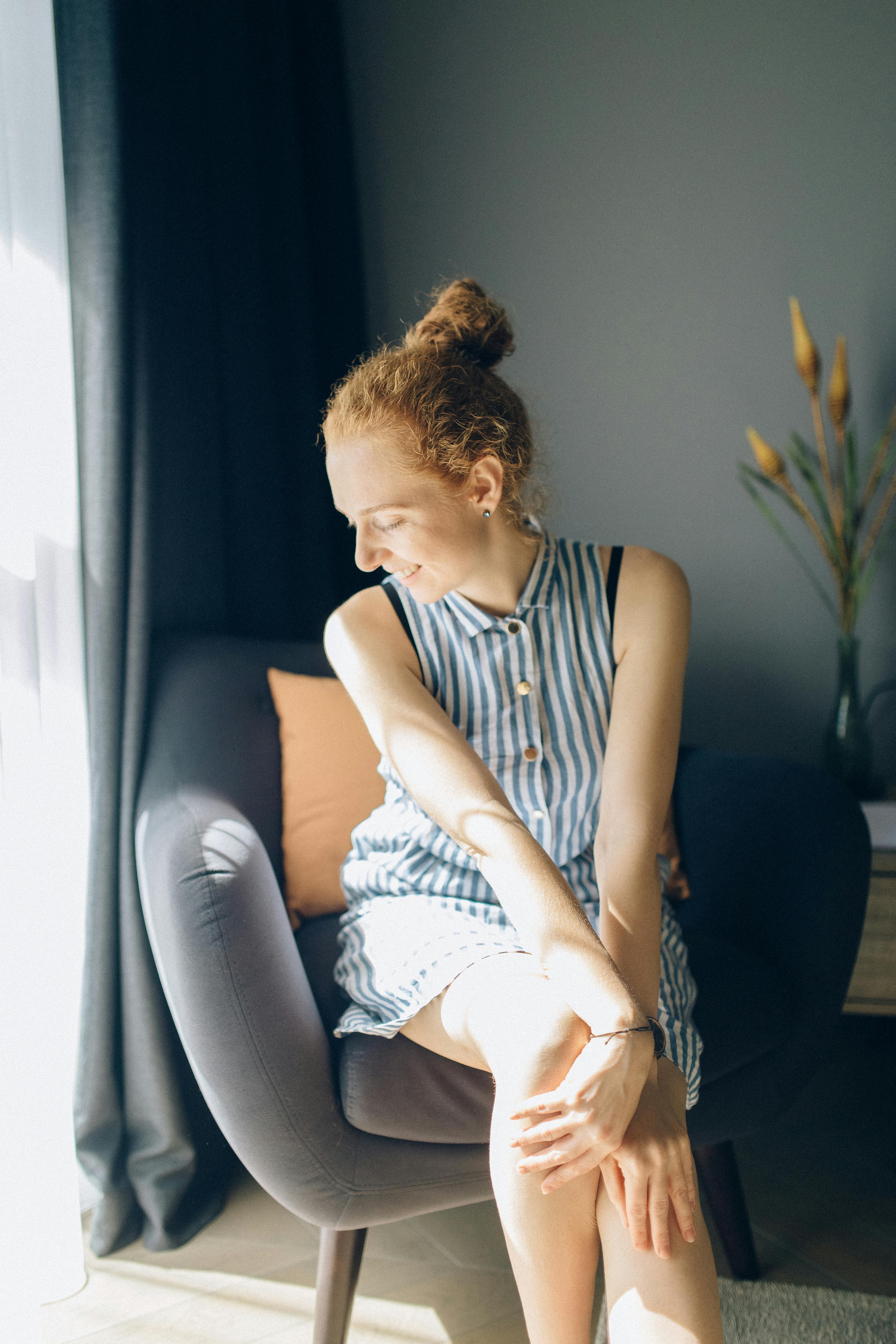 Redhead woman in a striped dress enjoys sunlight indoors, smiling warmly.