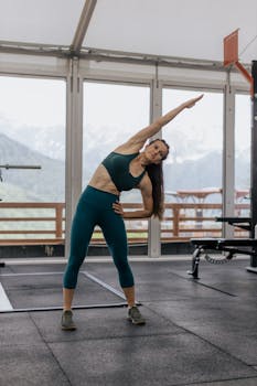 透过健身房窗户，可以看到一位白人妇女在做侧伸懒腰，远处是连绵的山脉