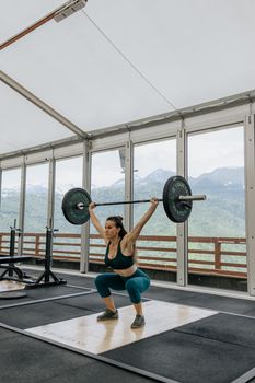 Adult woman lifting weights with mountains visible through gym windows.