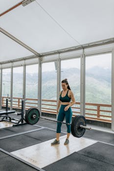 Adult woman lifting a barbell in a modern gym with scenic mountain views, showing strength and focus.