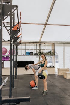 Two adults exercising with medicine balls in a modern gym setting.