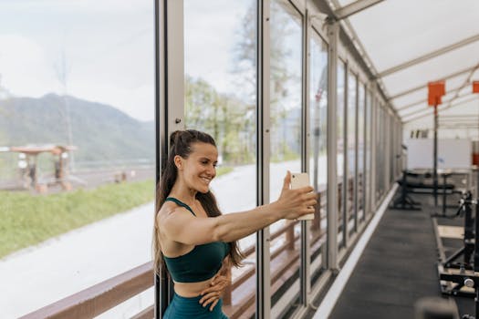 Smiling woman in sports bra takes a selfie inside a gym with large windows overlooking nature.