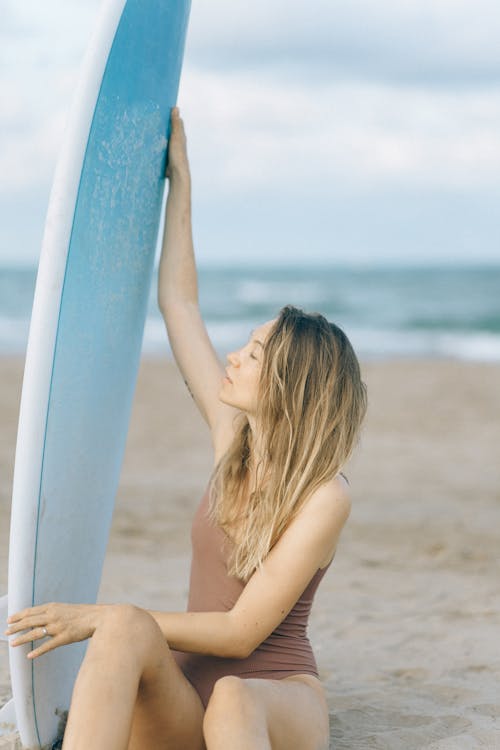 A Woman Sitting on the Sand and Holding Blue Surfboard