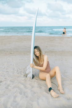 A young woman in swimwear sits on the sandy beach, holding a surfboard near the shore.