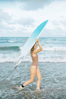 A woman in a swimsuit carries a surfboard along a sunny beach shoreline.