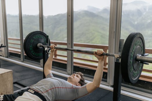 A man performs a bench press in a gym with mountain views, emphasizing strength and fitness.