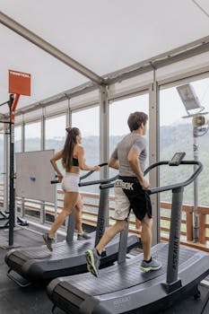 Man and woman running on treadmills in a bright gym with scenic views.