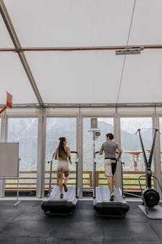 Back view of a couple running on treadmills in a gym with a scenic mountain view.