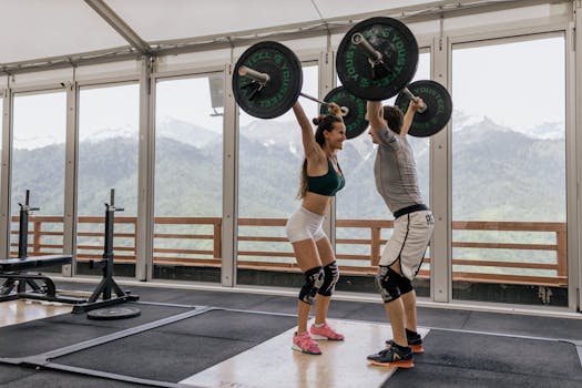 A couple lifting barbells together in a gym with scenic mountain views.