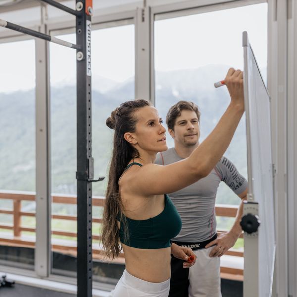 Female fitness coach writes on whiteboard as a male athlete watches in a gym.